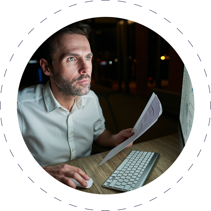 Man in white shirt working on computer, holding papers in dimly lit office.